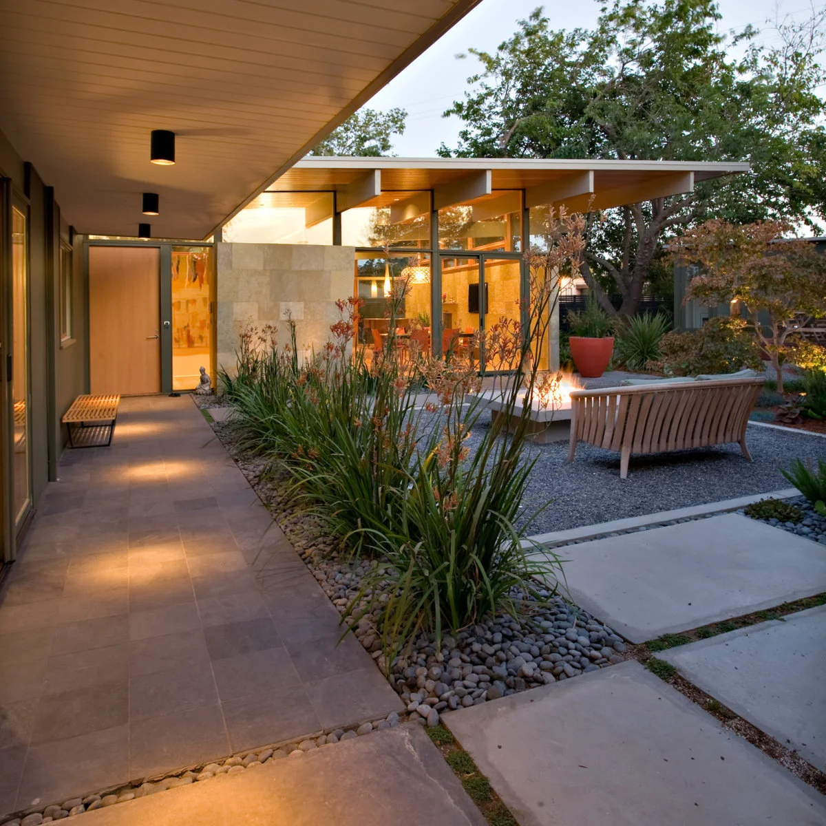 A broad pale overhang shelters a paved walkway and bench beside an open gravel seating area with planted borders.