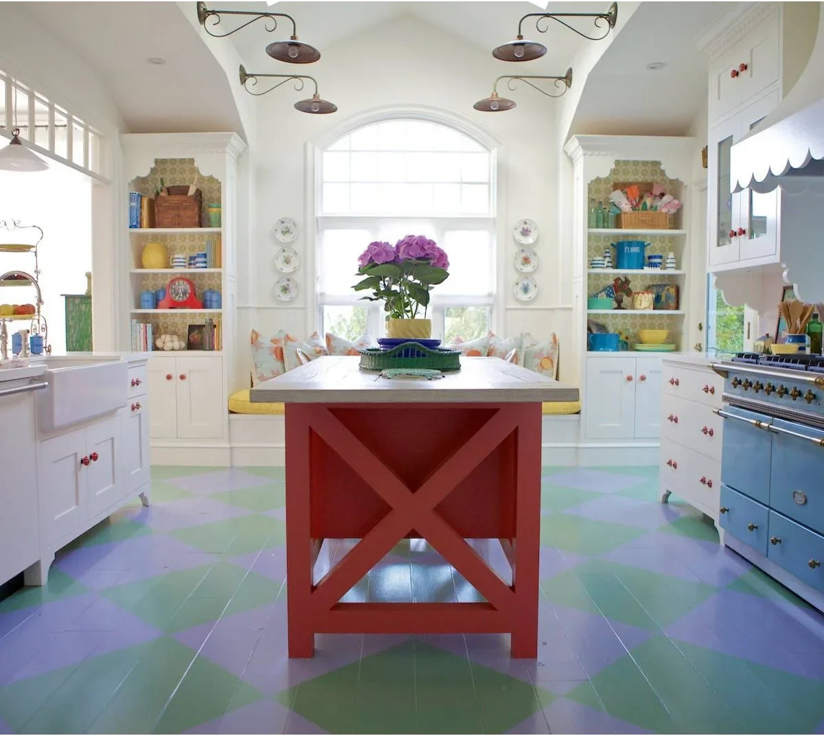 A red island stands in a white kitchen with a pale blue range, a green and lavender diamond-patterned floor and colorful open shelves.