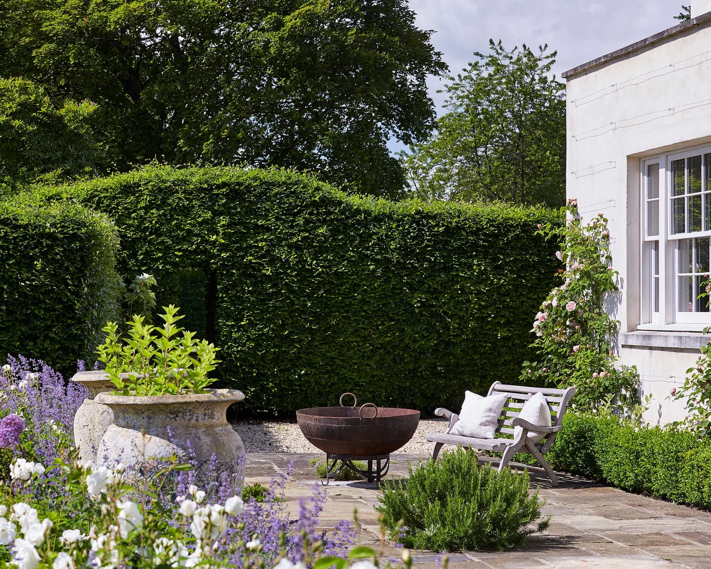 A pale garden bench sits beside a paved area enclosed by tall clipped hedges