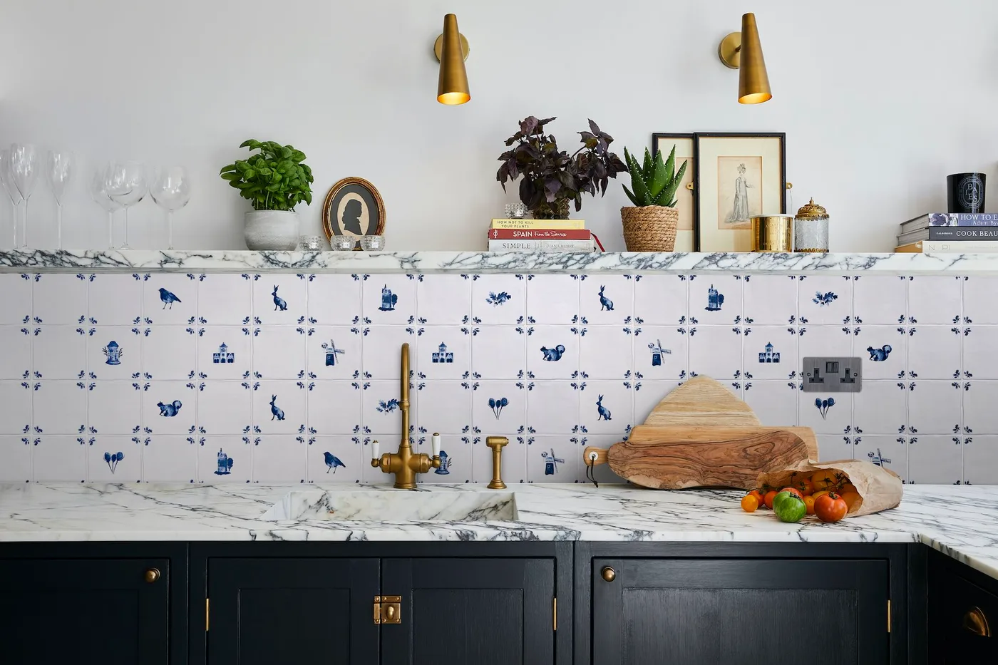 Navy lower cabinets sit below a pale veined counter and white tiles with small blue motifs beside warm-colored wall lights and a shelf