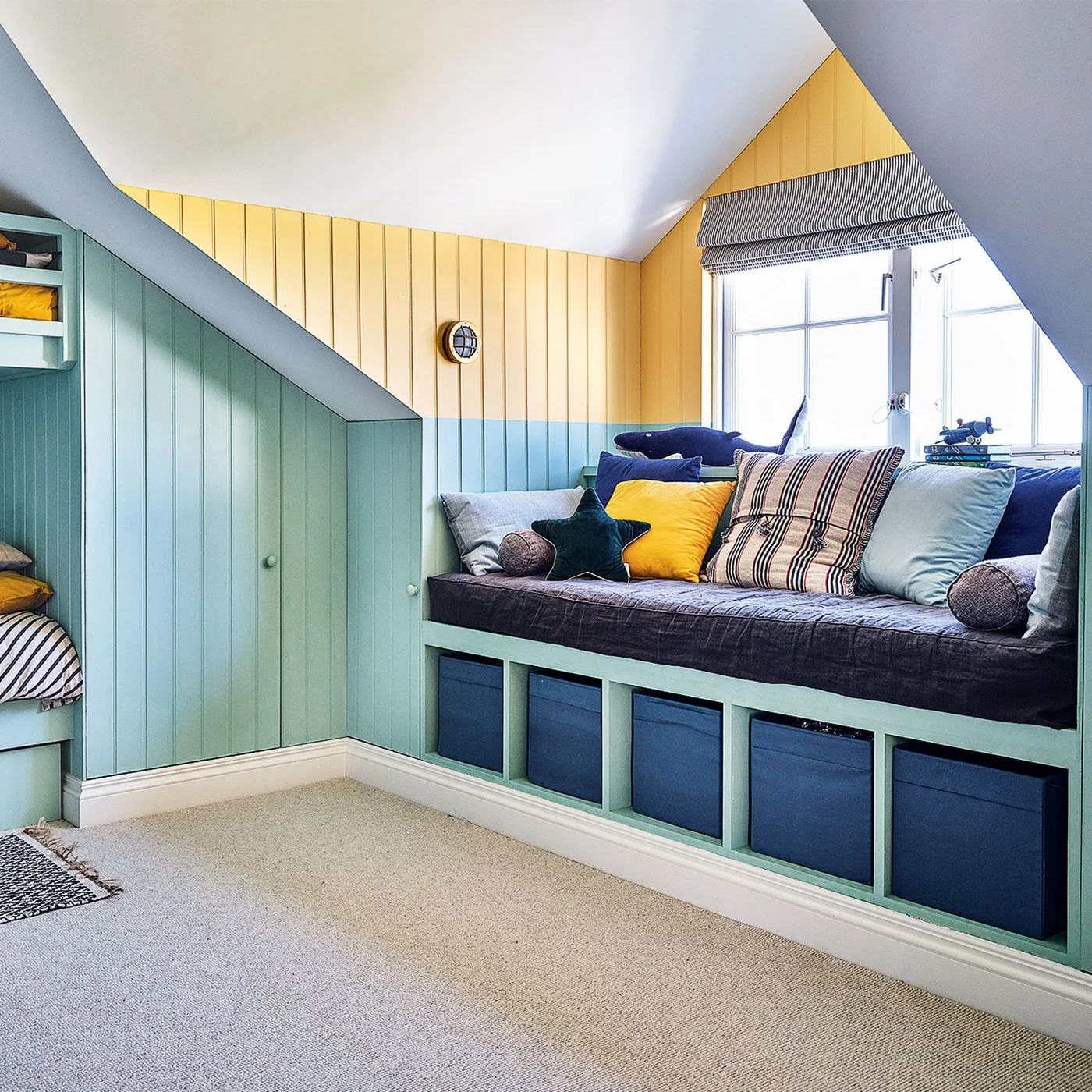 A window seat with dark cushioning sits above blue storage boxes beside two-tone green and yellow paneling in a sloped-ceiling children’s room