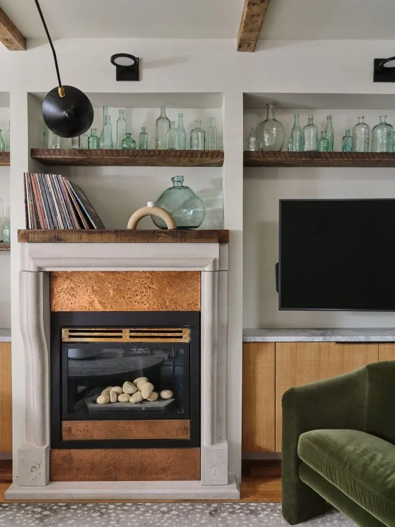 Wood shelves displaying pale glass bottles above fireplace and adjacent television recess with lower cabinet