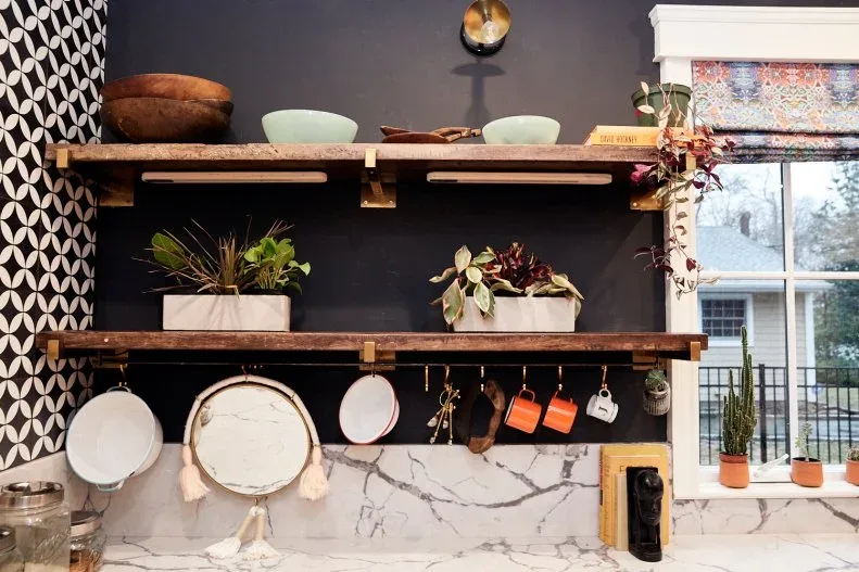 Dark kitchen wall with two wood shelves, pale bowls, plants, hanging mugs and a patterned blind.