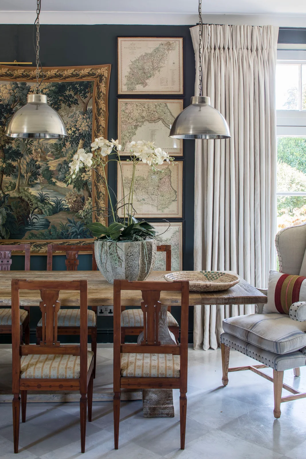 Framed maps and a large scenic tapestry hang on a dark blue dining-room wall behind a wooden table beneath two silver-colored pendants