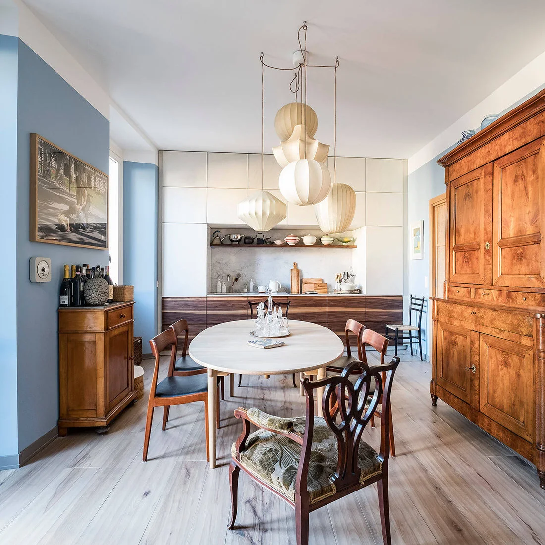 An oval pale dining table and mixed wood chairs stand between older wood cabinets and a modern kitchen beneath sculptural pale shades.