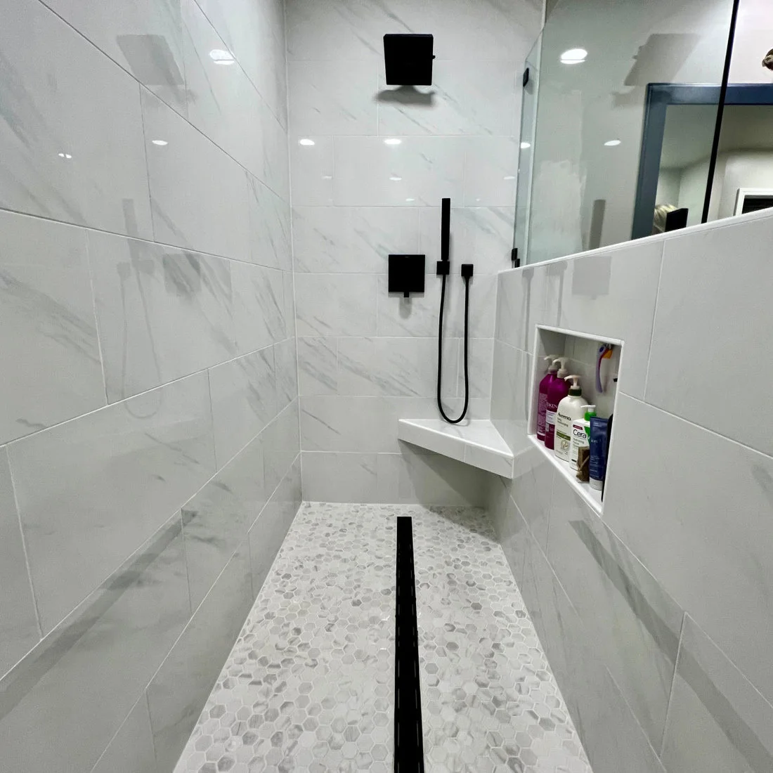Small pale hexagonal mosaics surround a black linear shower drain beneath large white wall tiles.