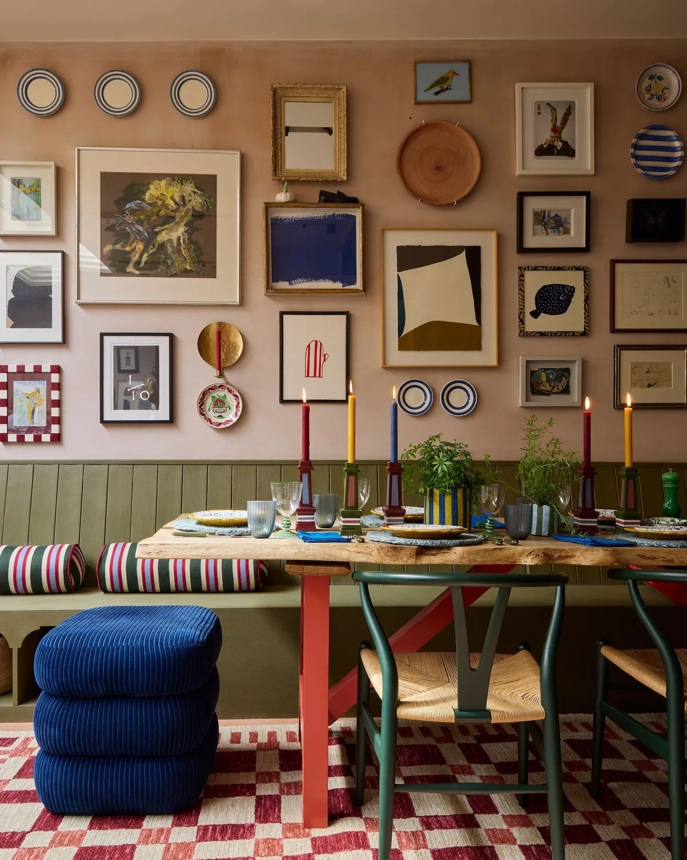 Dining room with an olive bench, mixed framed art and plates, a wood table with red legs, green chairs and a checked rug