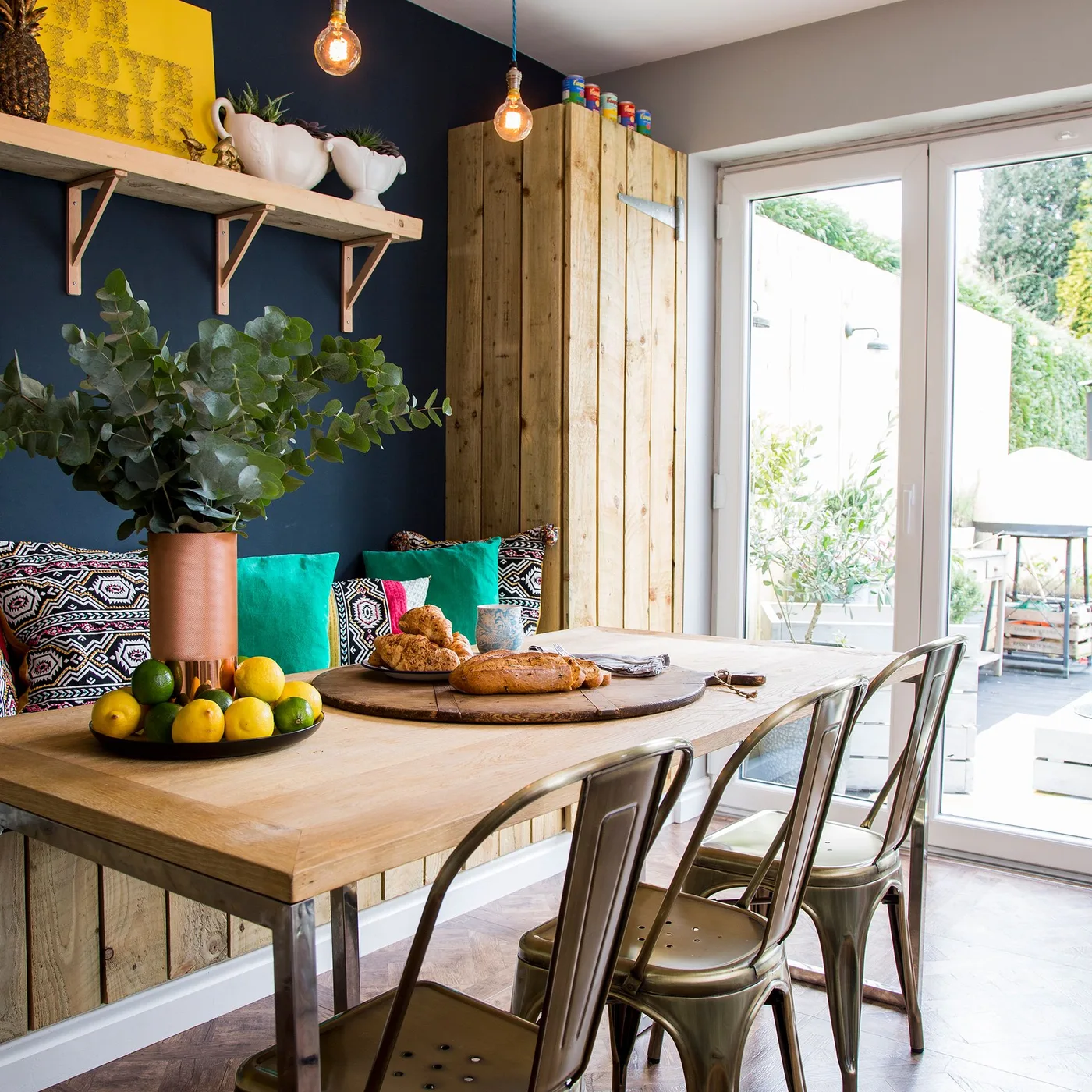 A wood dining table and metal chairs face a cushioned bench beneath a dark blue wall beside glazed doors.