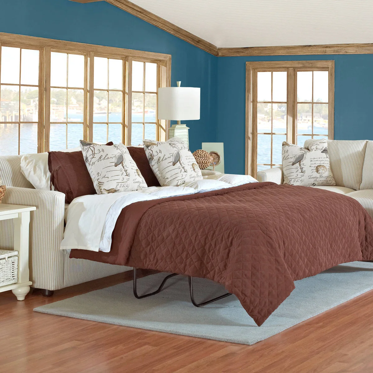 Striped sofa bed opened with white sheets and a brown quilt beside a white side table