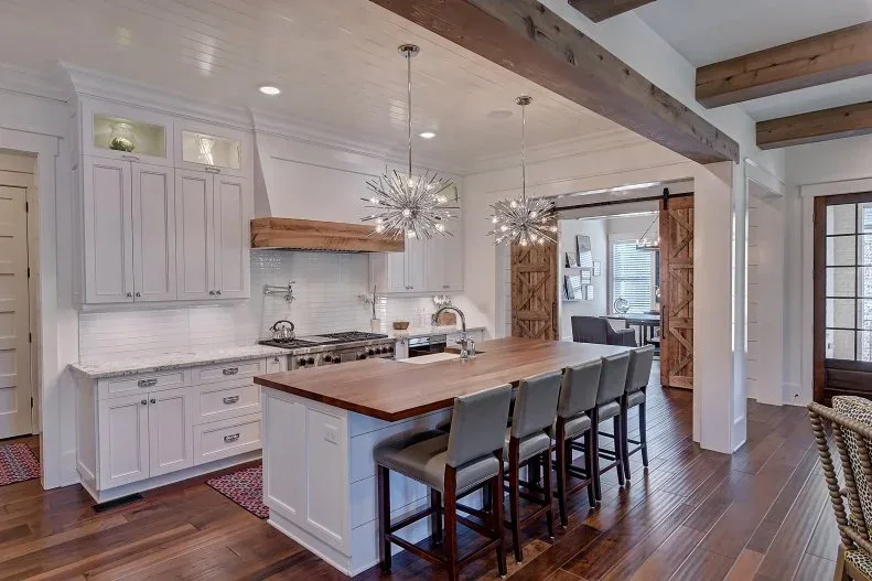 White farmhouse kitchen with wood-topped island, upholstered stools, exposed beams, spiky silver pendants and a sliding wood door