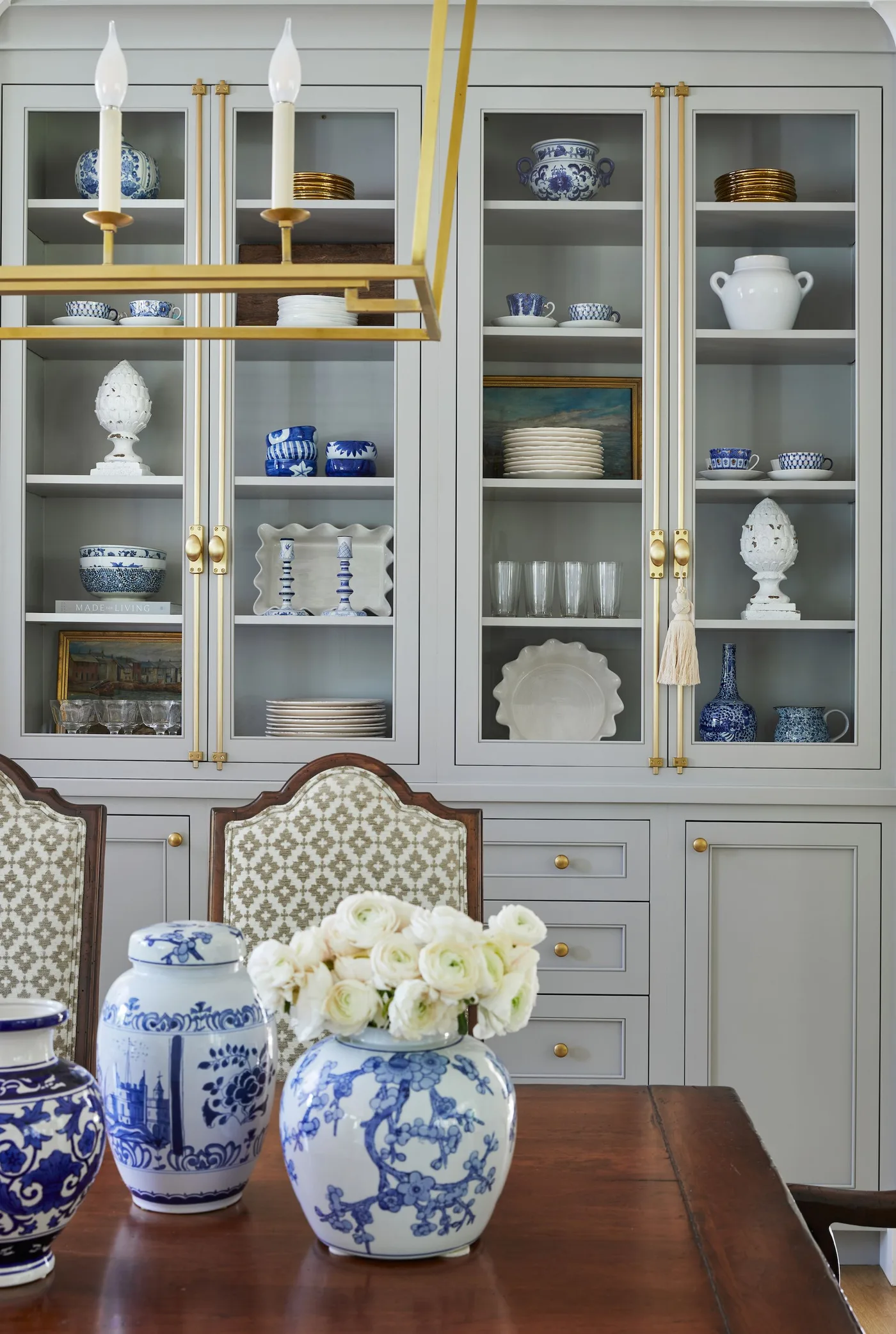 Gray glass-fronted cupboard with blue-and-white dishes behind a wooden table, patterned chairs and a gold-toned light.