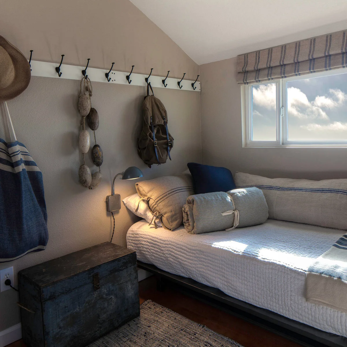 Small sleeping nook with a striped single bed, wall reading light, blue cushions and a dark trunk