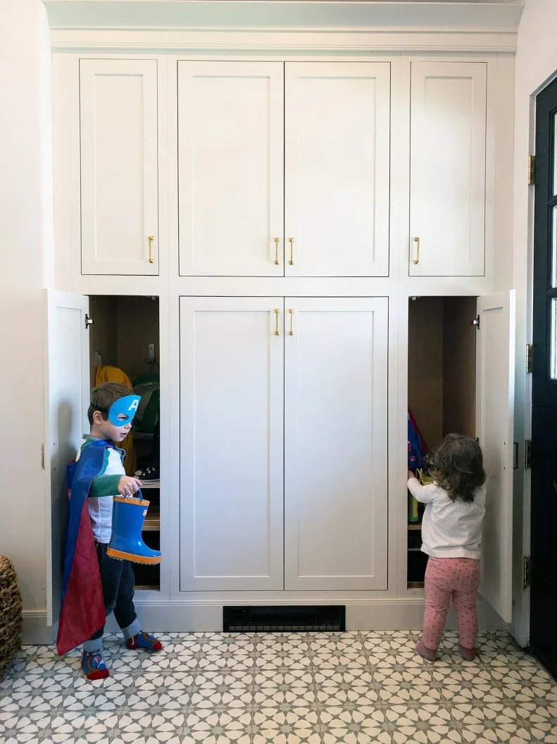 A white floor-to-ceiling mudroom cabinet has open lower side compartments, closed upper doors and brass-colored handles above a patterned floor.