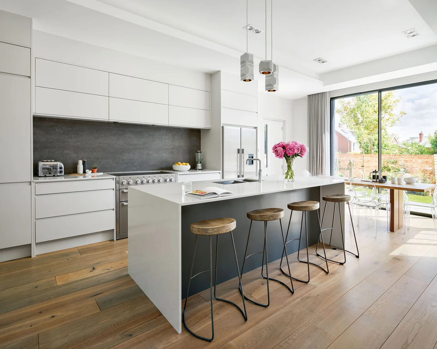 White flat-front kitchen cupboards and a gray backsplash stand behind an island with a dark seating face and four wood-topped stools