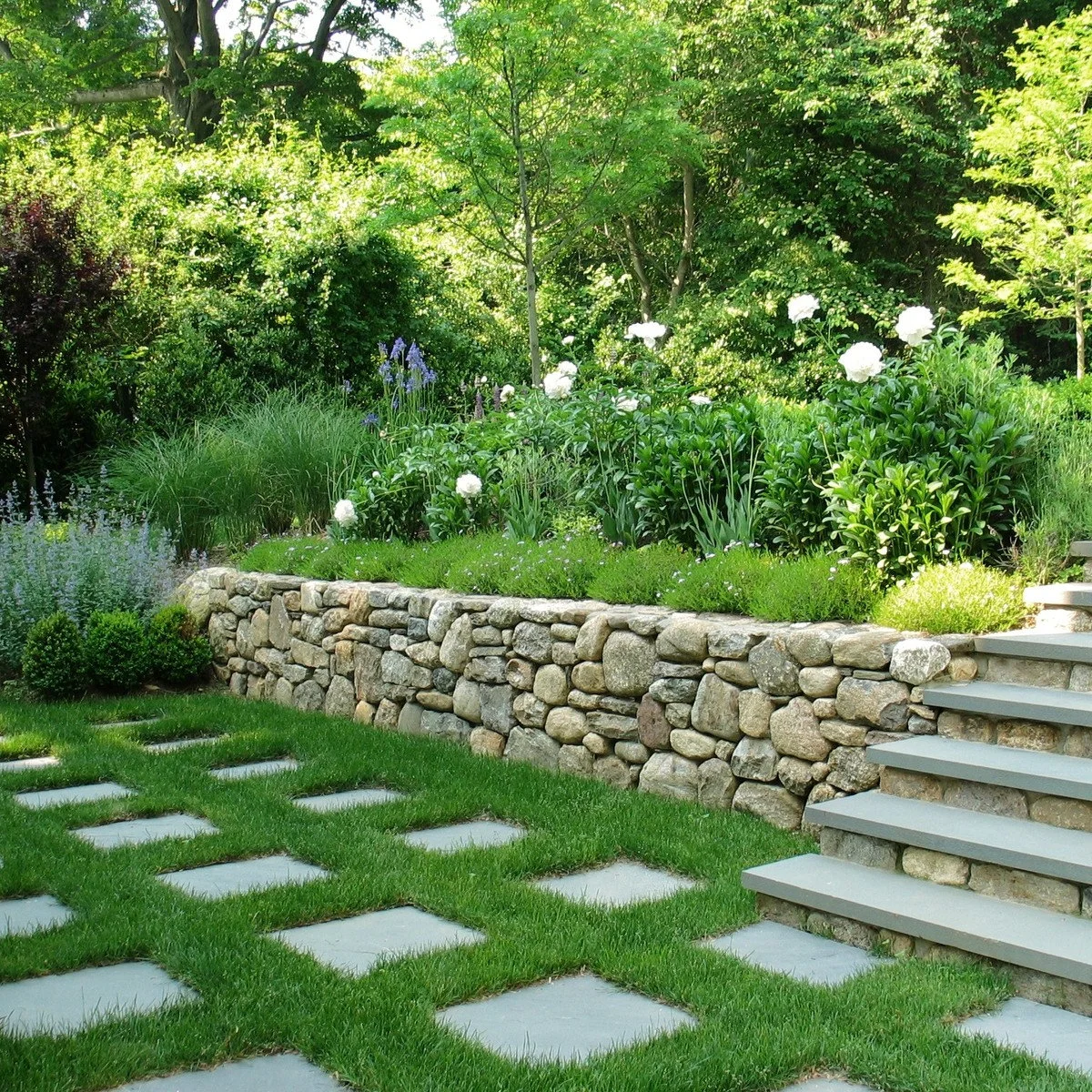 An irregular stone retaining wall with flowering planting beside broad pale garden steps and lawn.