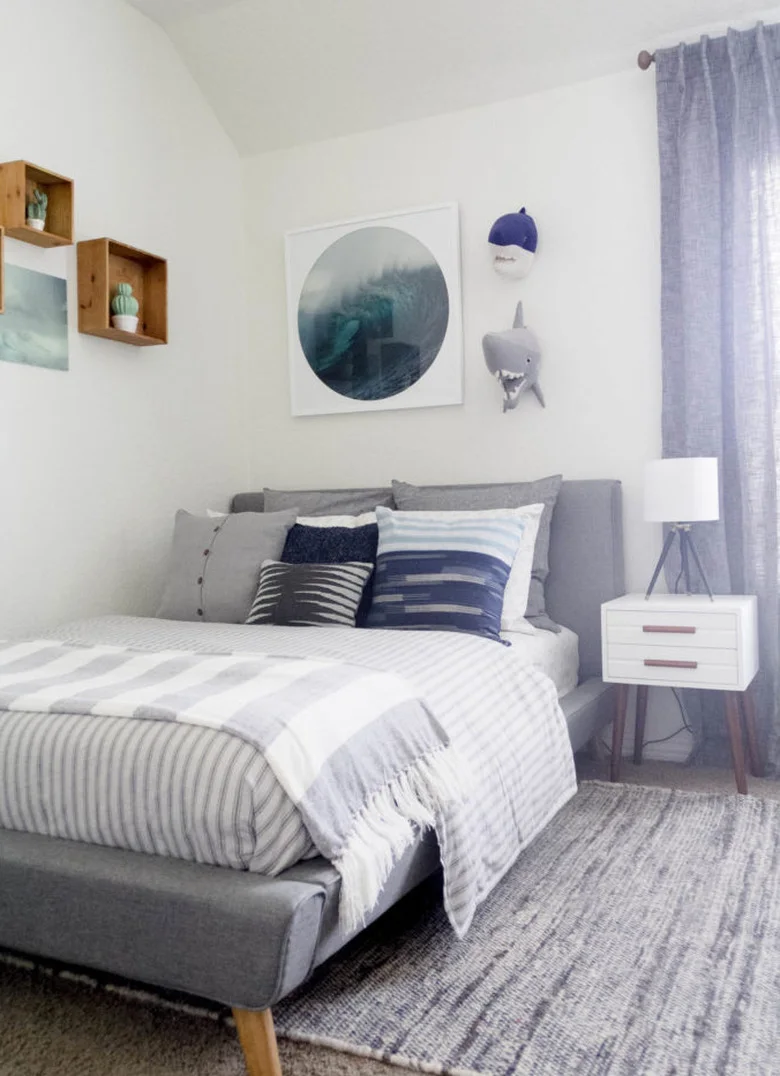 A gray upholstered bed has blue and gray striped pillows beneath ocean-themed artwork and two animal wall decorations beside pale walls and a white bedside unit