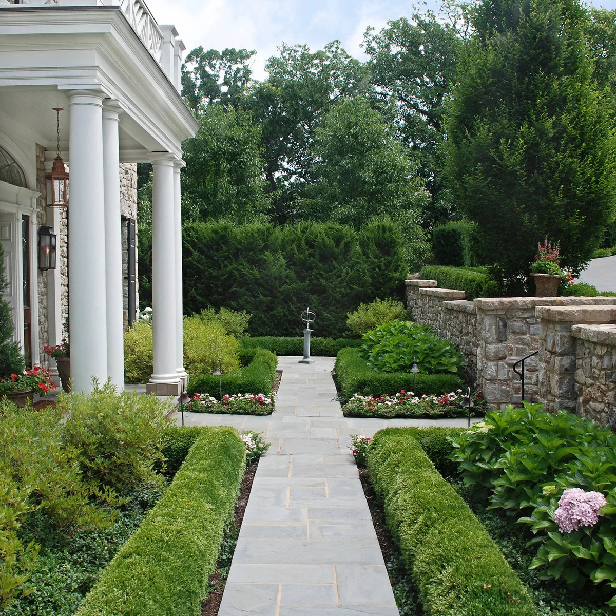 Low rectangular hedges outline stone paths and flower beds between a white-columned house and a stone boundary wall.