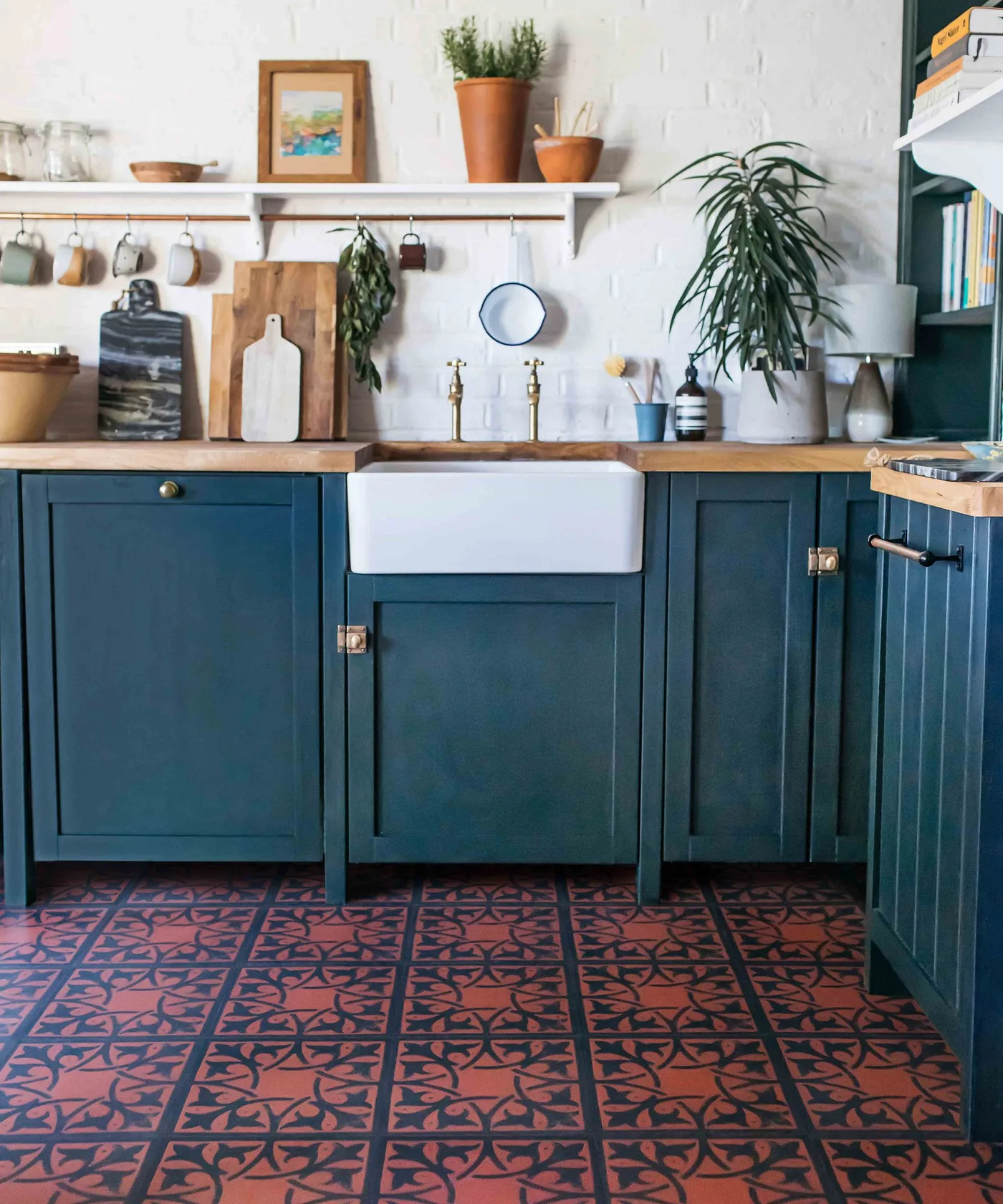 Kitchen with deep blue lower cabinets, white apron-front sink, wood counter, brass-colored taps, open shelf with mugs, and pink-blue patterned floor.