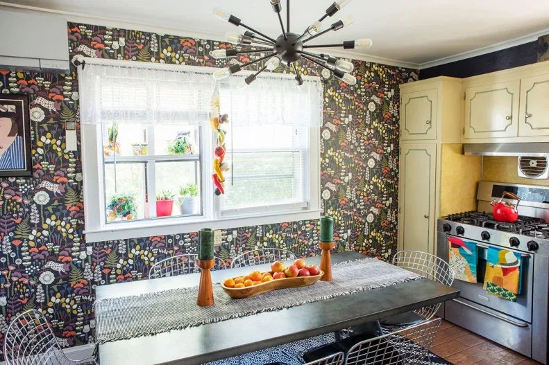 Eat-in kitchen with dark floral wallpaper, pale yellow cabinets, a dark table and wire chairs.
