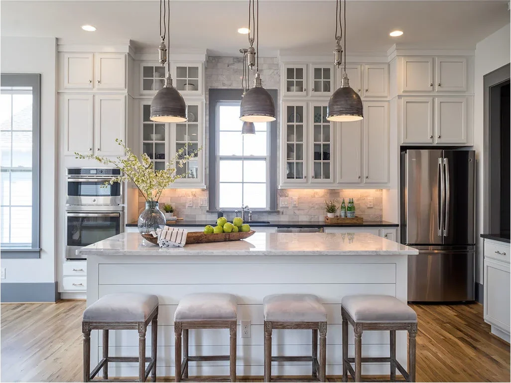 Two tiers of white cupboard doors rise above a stainless refrigerator in a kitchen with tall display cabinets.