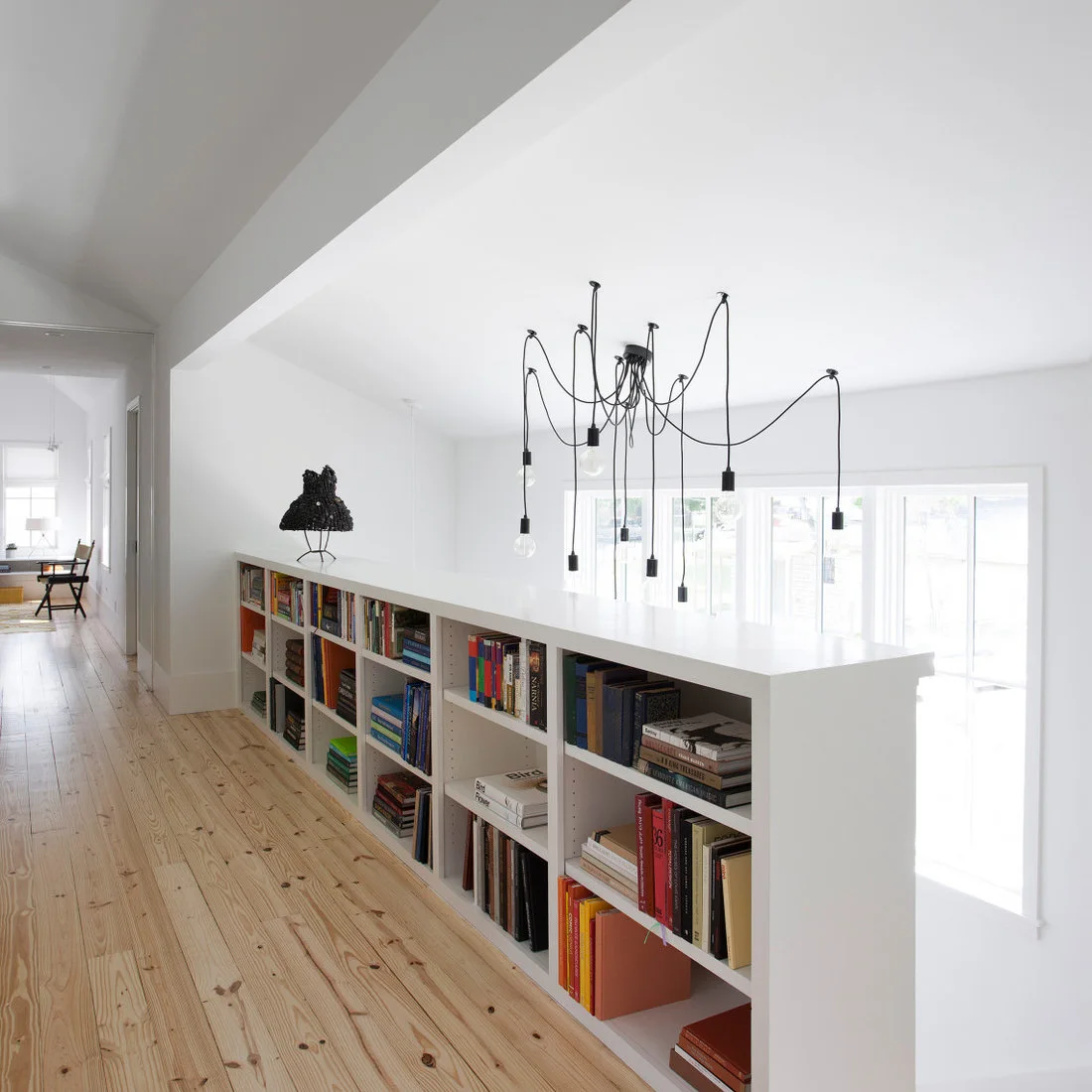 A long low white bookcase borders an upper hallway with pale wood flooring beside an open stairwell and hanging black lights.