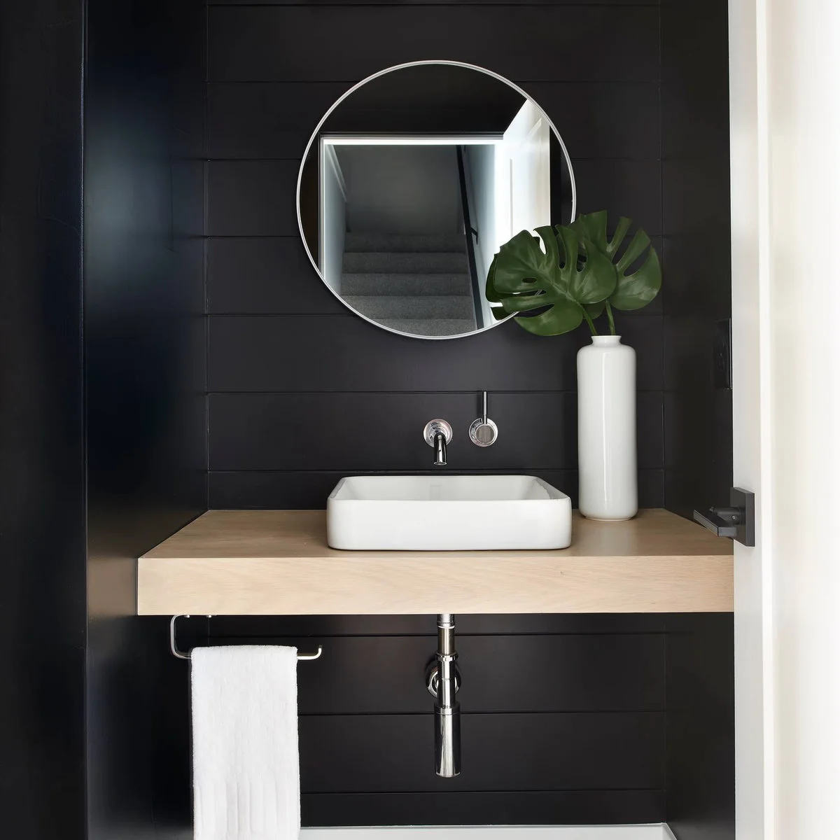 A white hand towel hangs from an open-ended rail below a wood counter with a rectangular vessel sink and dark paneled walls.