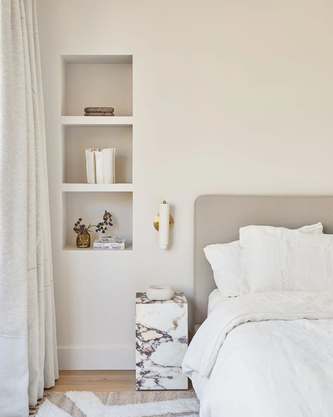 A pale upholstered bed stands beside three recessed display niches, a cylindrical wall light, a veined rectangular bedside block and long light curtains.