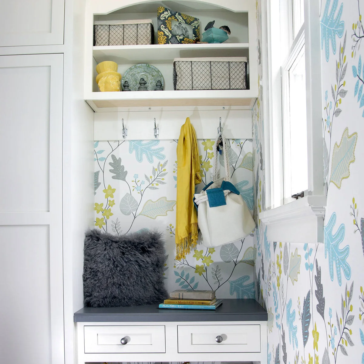 Small fitted entry seat with gray top, white drawers, wall hooks and blue and yellow patterned wallpaper.