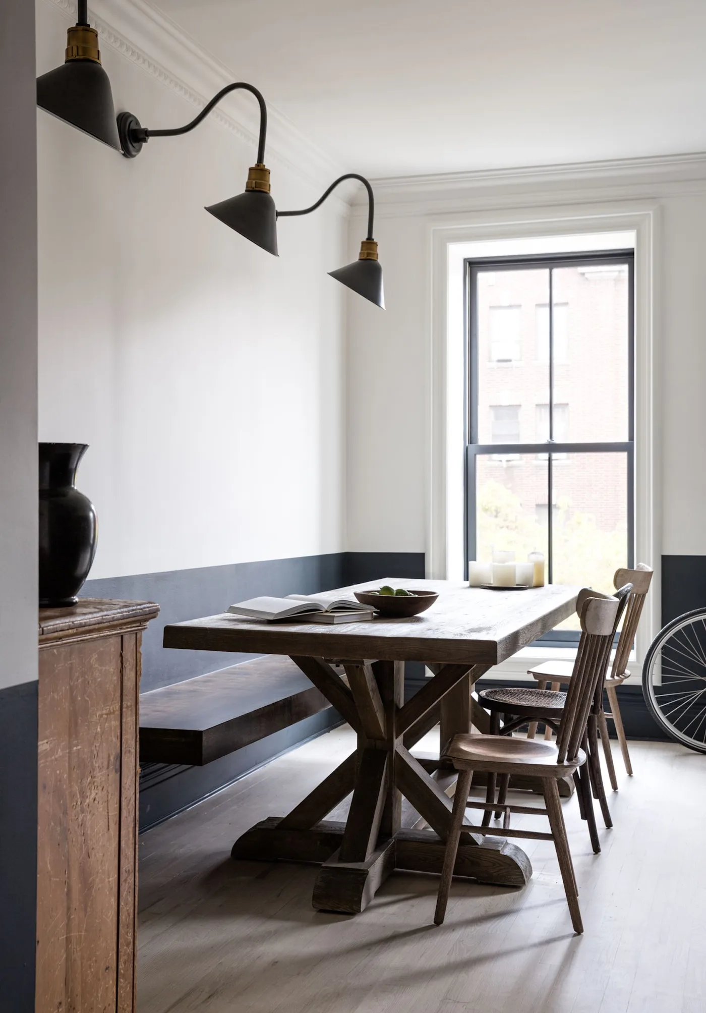 Country kitchen dining corner with long wooden table, straight bench, wooden chairs, dark lower wall and three dark wall lamps.
