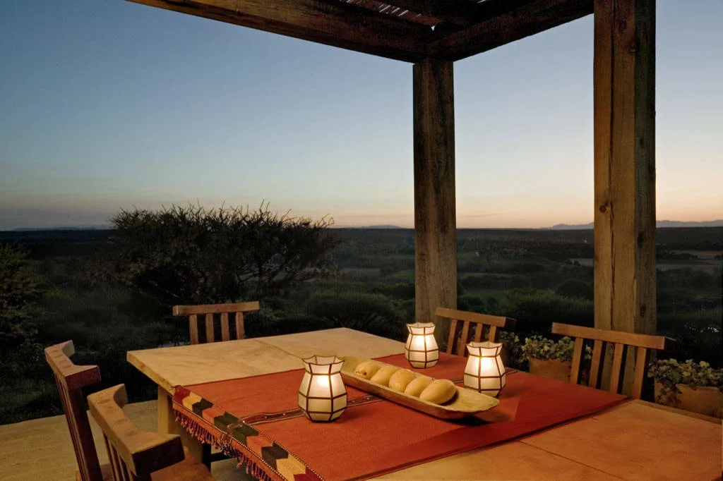 Wood outdoor dining table with an orange runner, three small lanterns and a low bowl of fruit under a shaded roof.