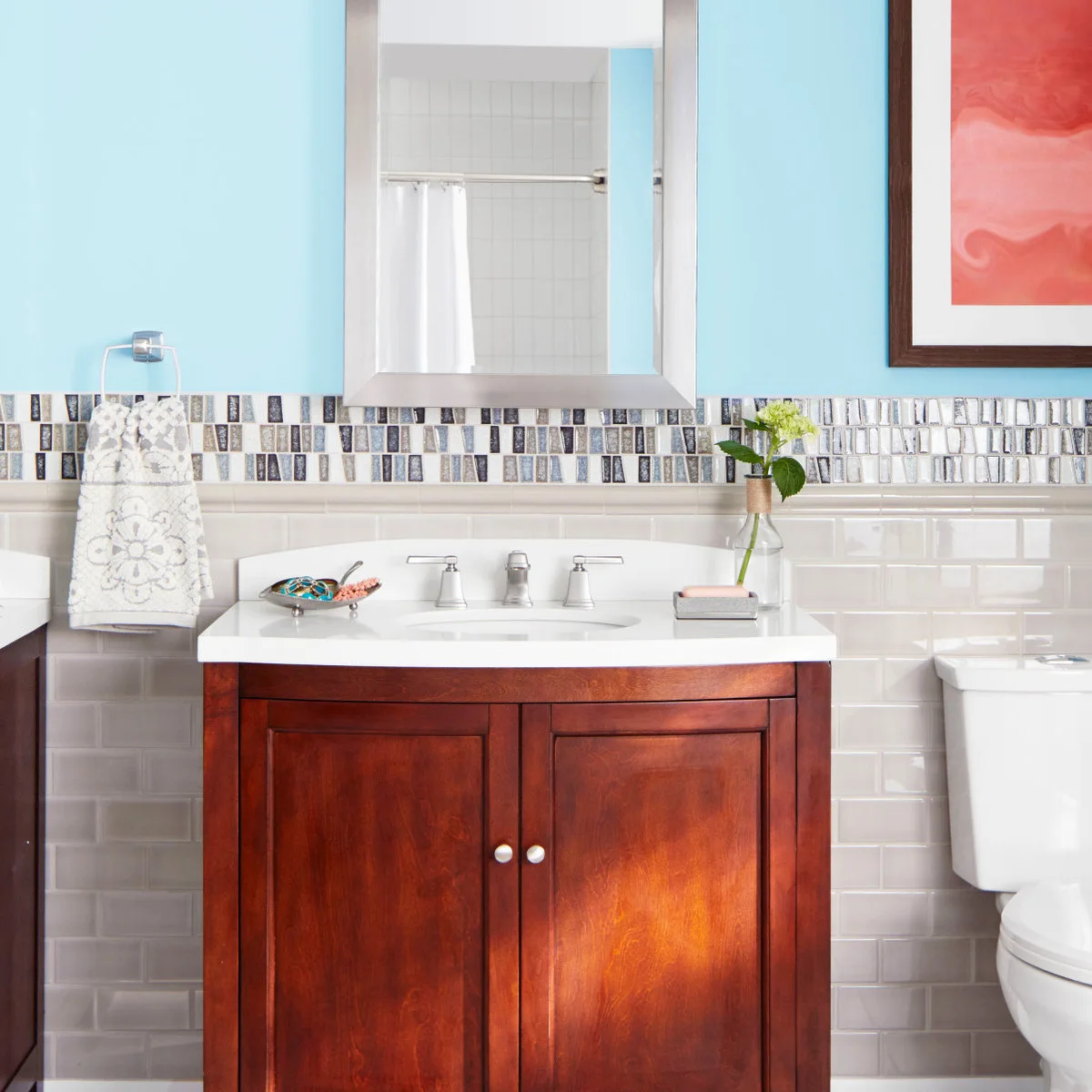 Blue painted wall above pale subway tile and a mosaic border around a wood vanity and framed mirror.
