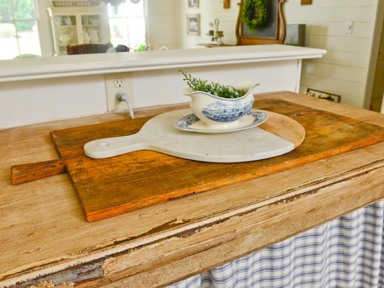A worn timber cabinet top holds layered boards and a blue-and-white jug above a checked fabric curtain.