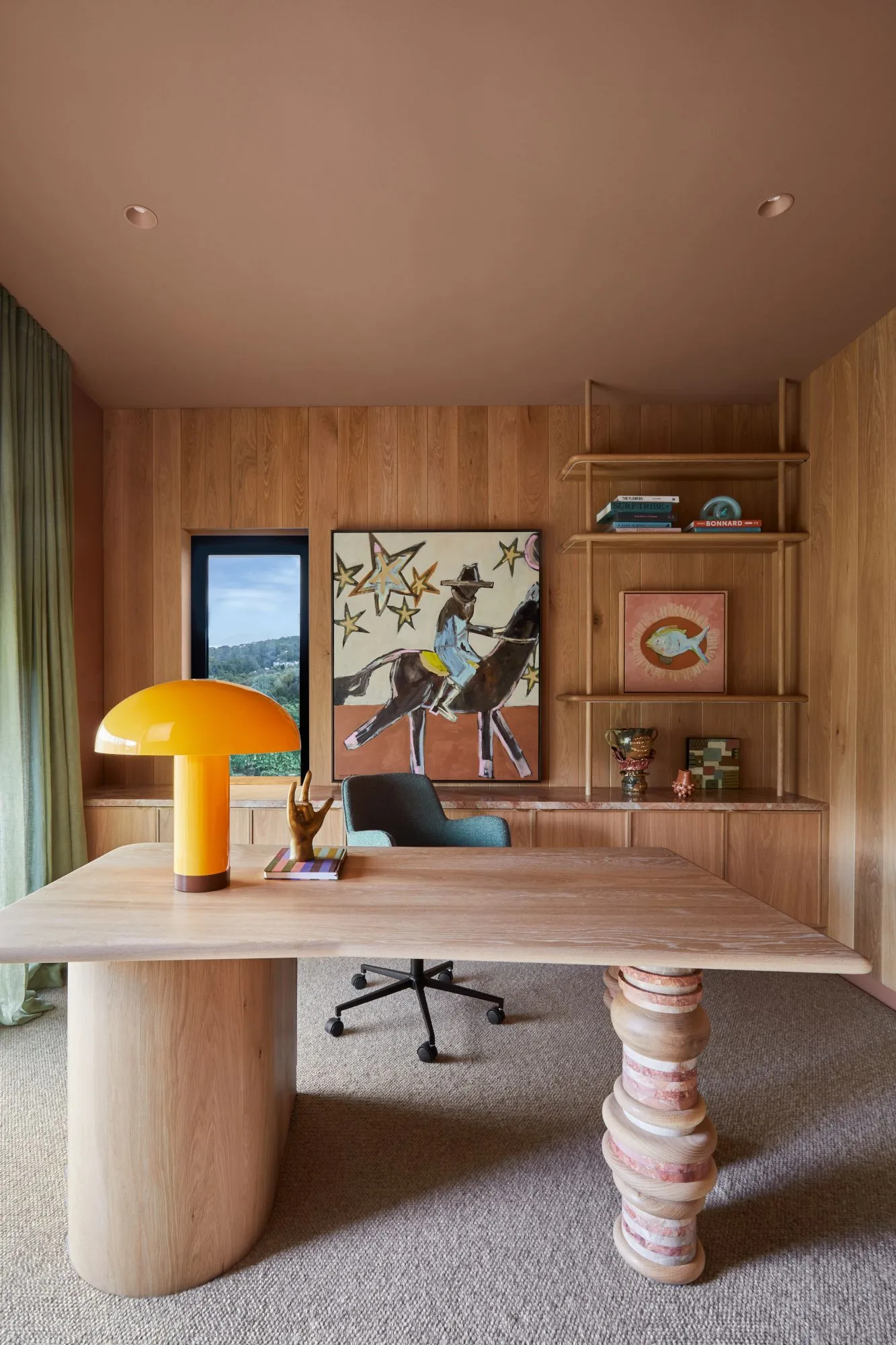 Home office with wooden paneled walls, pink ceiling, broad desk with differently shaped supports, yellow dome lamp, framed artwork and teal rolling chair.