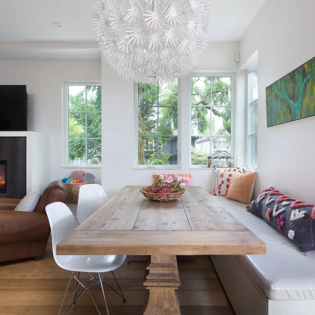 A wood dining table stands beside a pale corner bench with colorful cushions, next to a brown sofa and a wall television at the room’s edge.