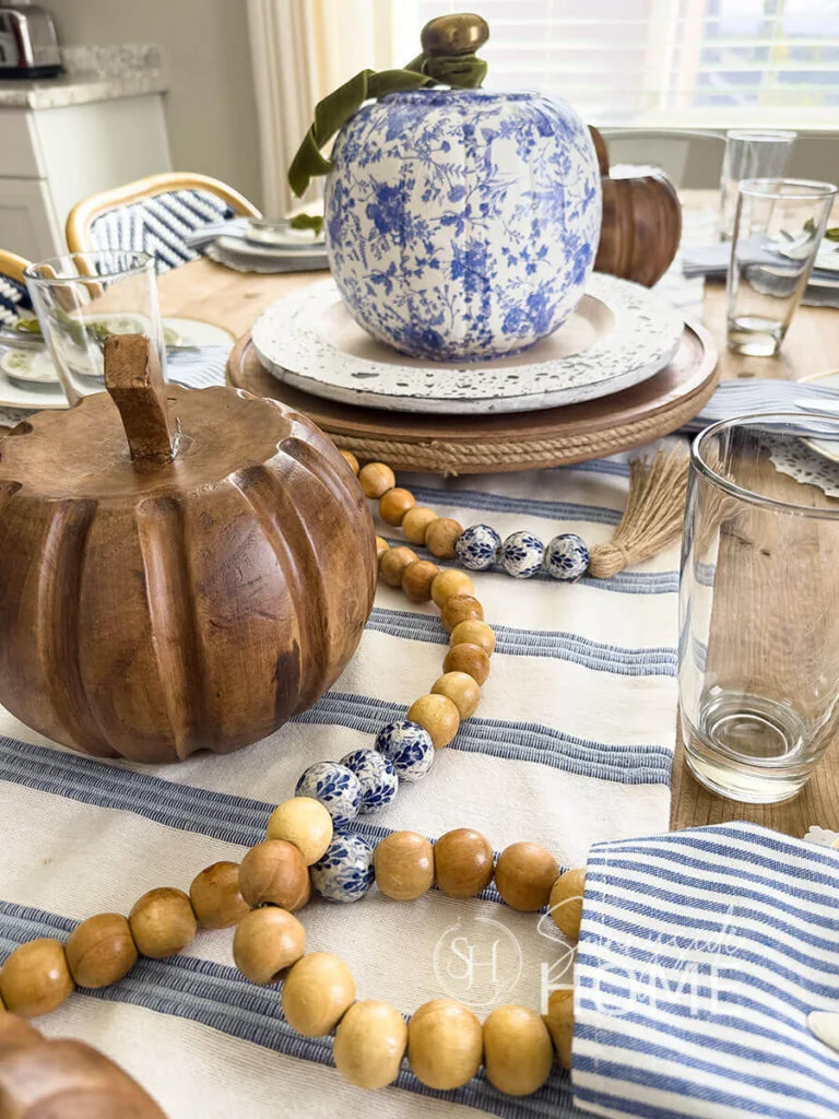 A blue-and-white patterned pumpkin with a green ribbon sits above a round stand beside smaller wood-colored pumpkins and a bead garland