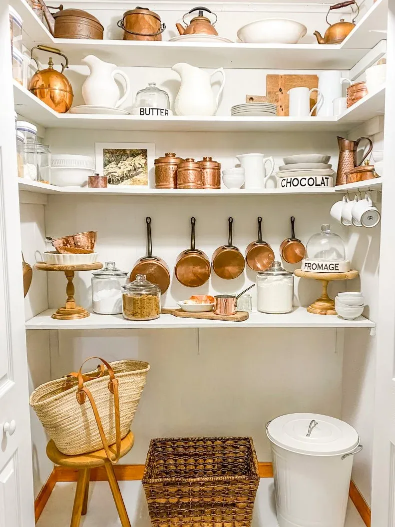 White country pantry shelves with copper-toned pots, pale pitchers, glass jars, baskets and hanging cookware