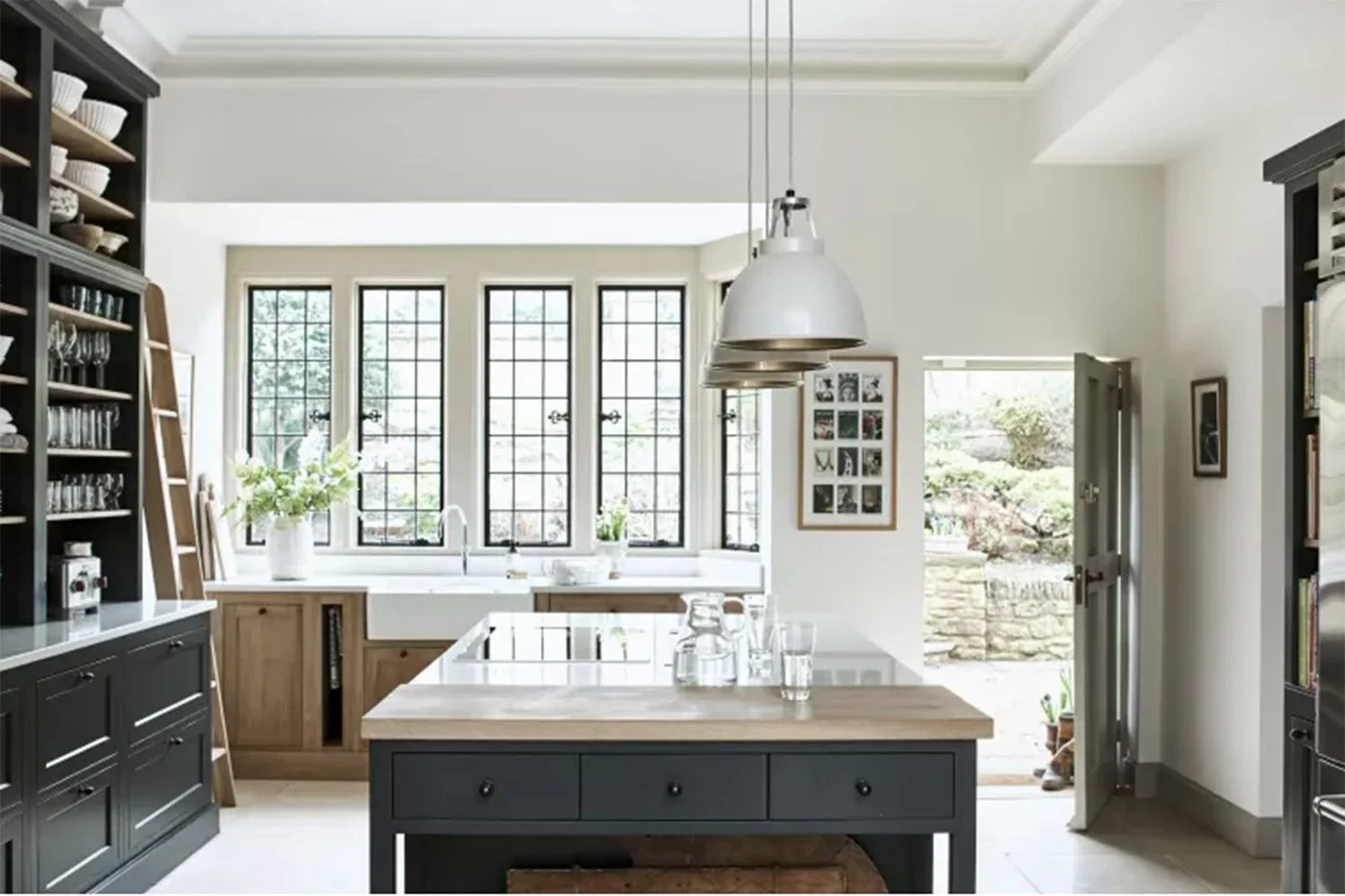 A light kitchen has dark glazed storage and an island below pale upper walls beside black-framed windows, a white sink and warmer lower cupboards
