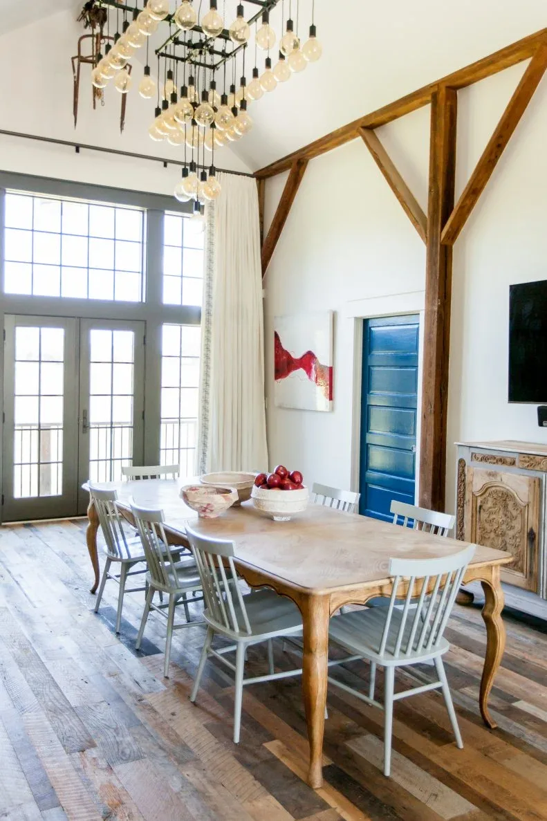 Light curved-leg dining table with gray spindle-back chairs, exposed beams and a vivid blue door.