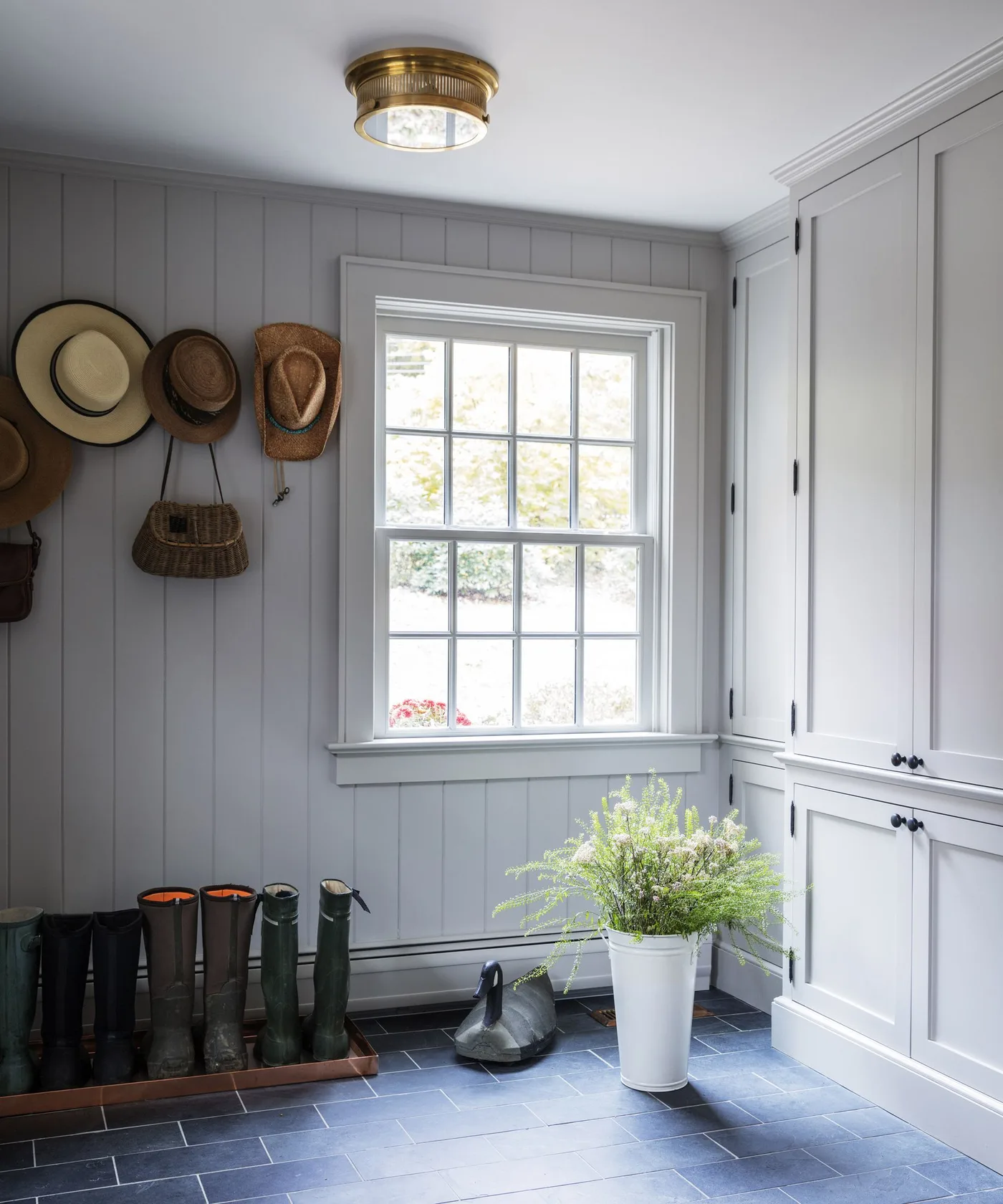 Light gray paneled entry with hats on wall hooks, tall boots on a low tray and closed cupboards beside a window