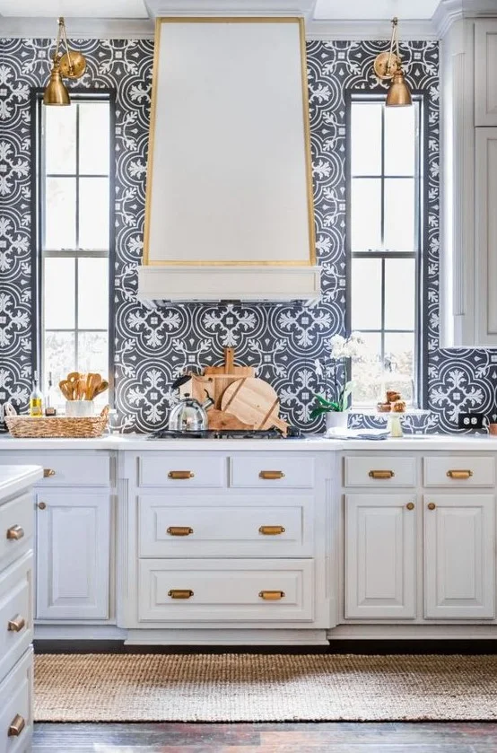 A pale hood with warm-colored edging stands between tall windows above blue-and-white patterned tiles, pale drawers and warm-colored hanging lights