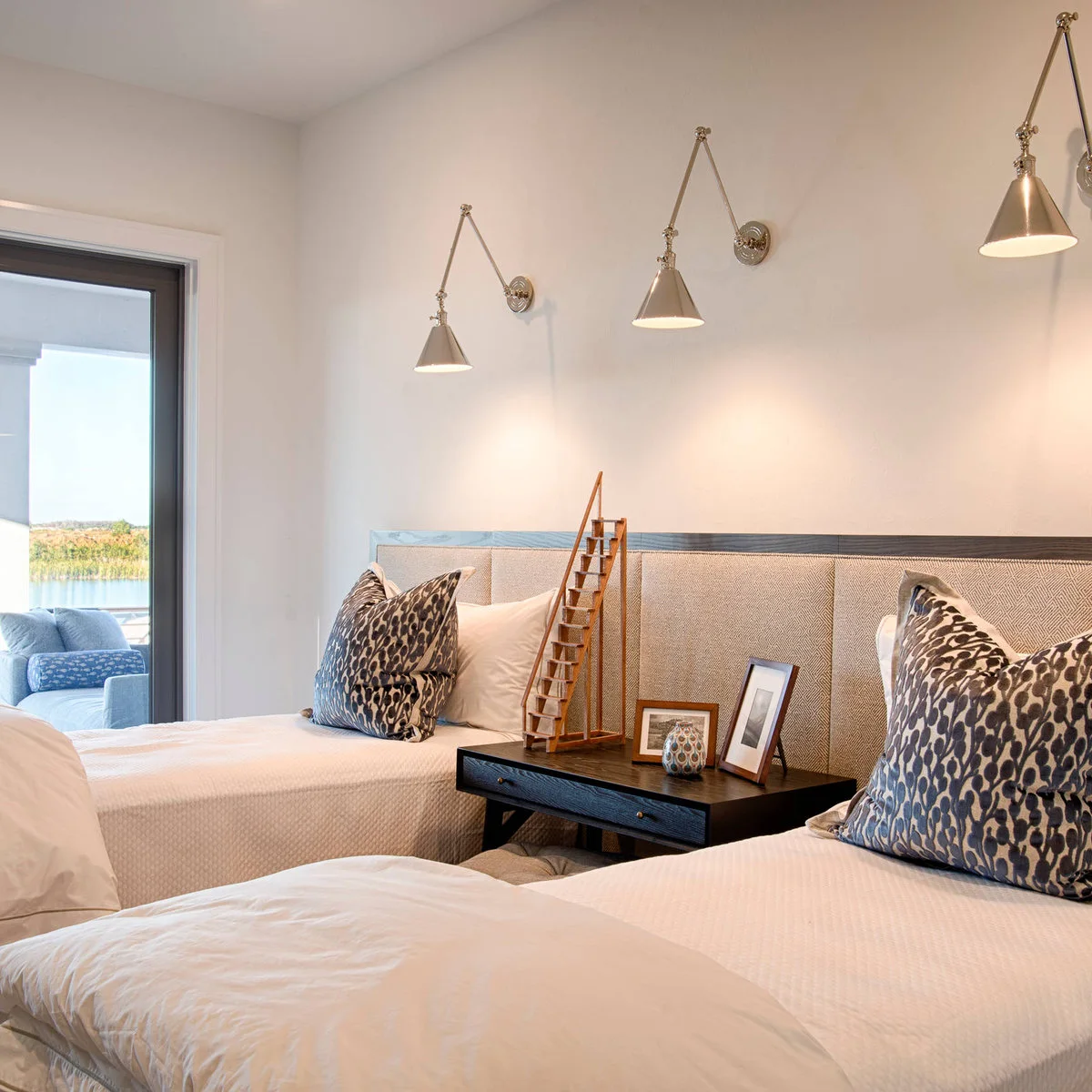 Two pale beds with a continuous neutral headboard, patterned cushions, shared dark table and wall-mounted lights