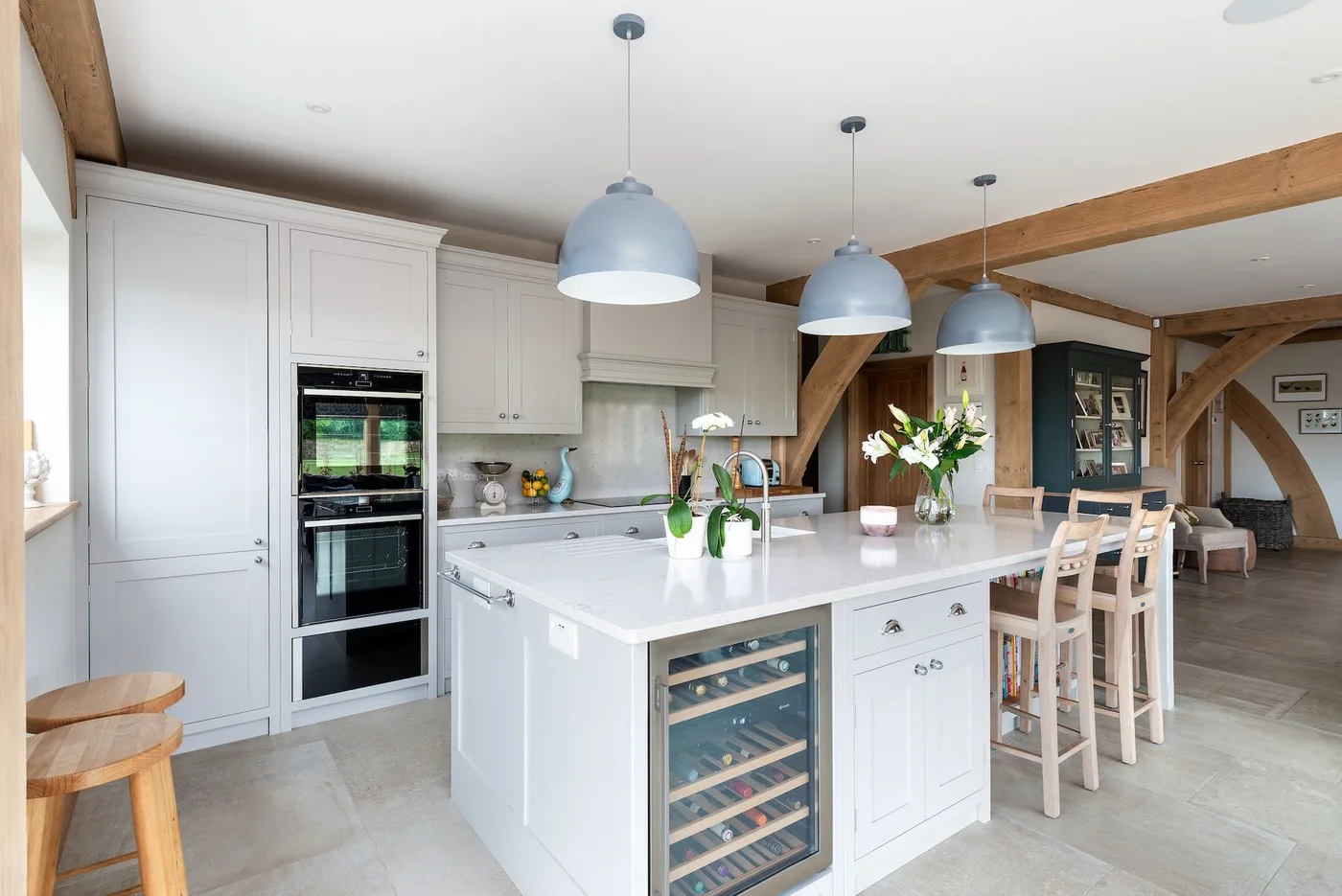 White kitchen with pale blue dome pendants, island stools, fitted ovens and exposed wooden beams.