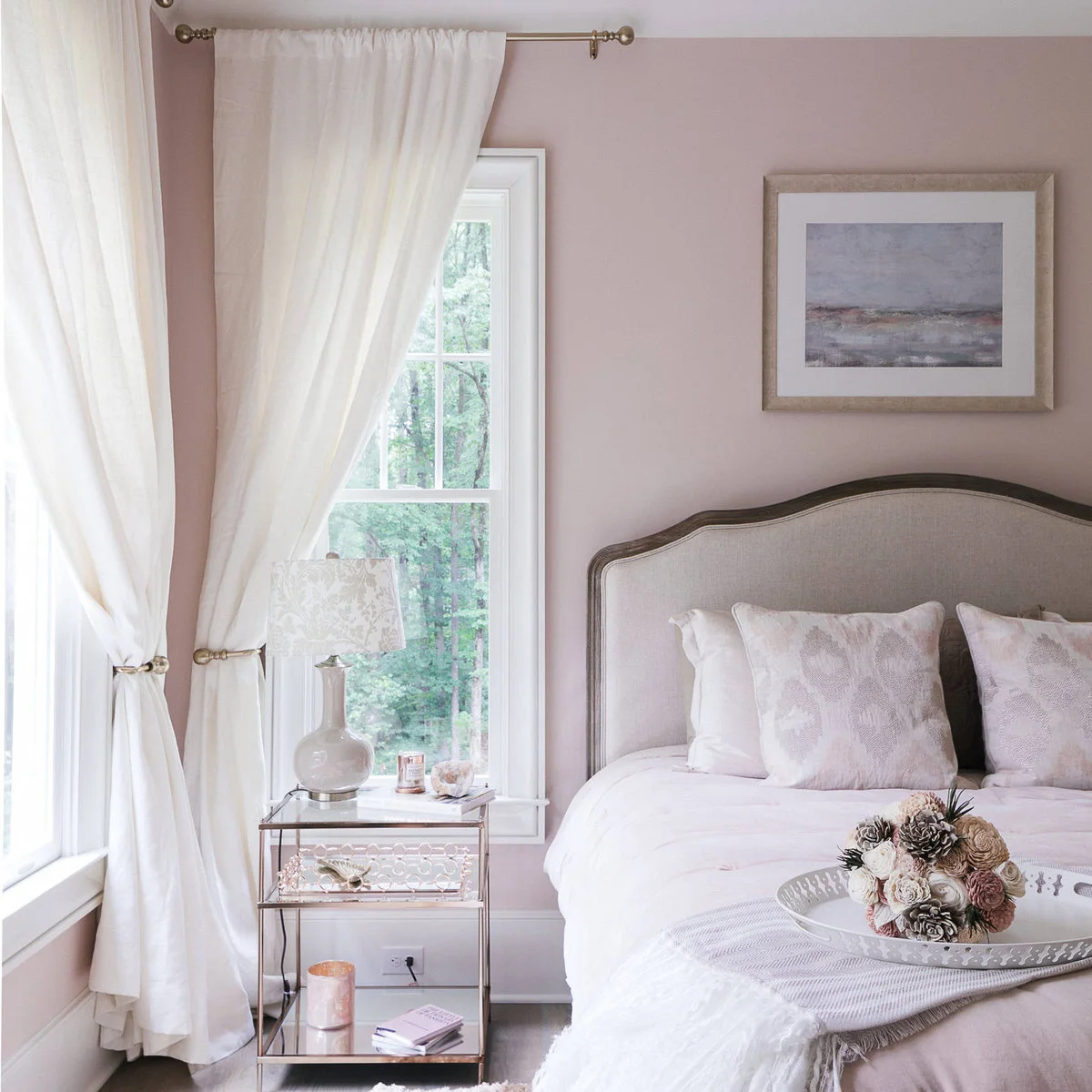 Pale blush bedroom walls with white curtains, a neutral upholstered headboard and light patterned pillows.