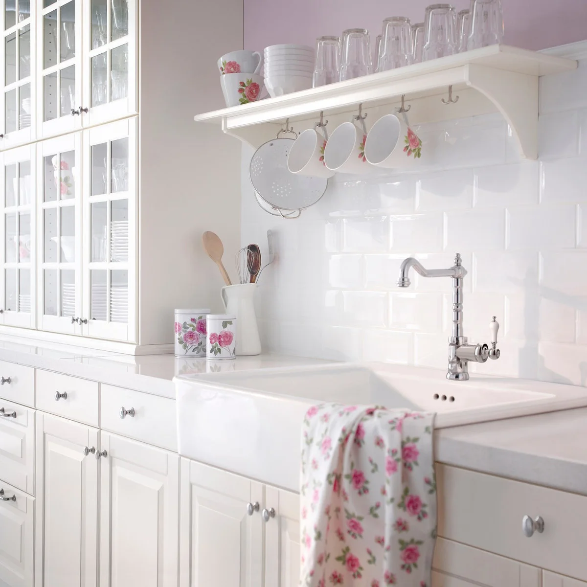 A white shelf with hooks and floral mugs above a white kitchen sink and tiled backsplash.