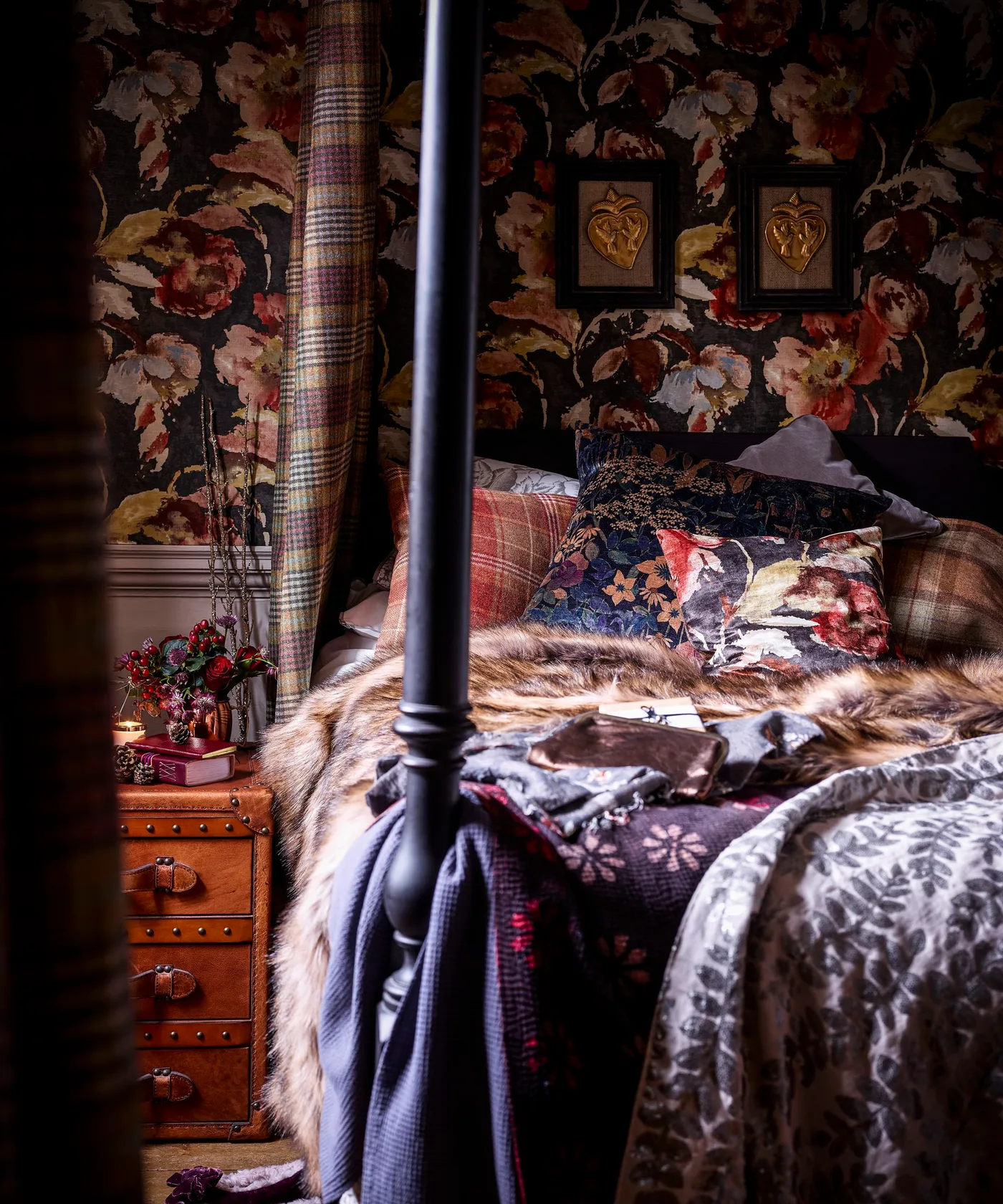 Dark floral bedroom wallpaper and four-poster bed with checked and floral cushions, shaggy throw, and a trunk-style bedside table.