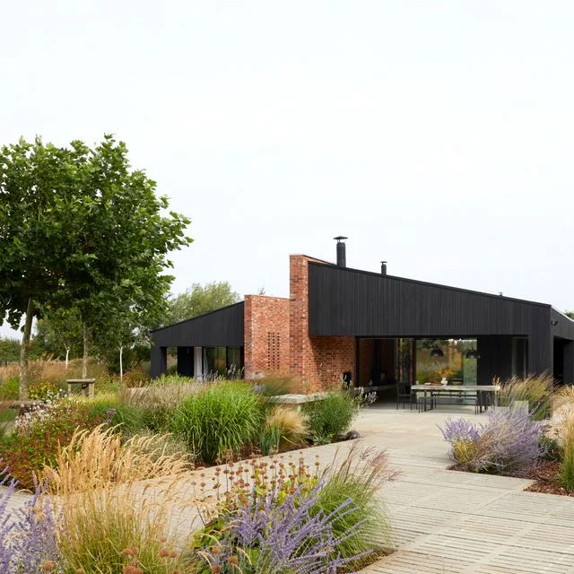 A pale paved route flows between flowering borders into a dining terrace beside a dark-clad house