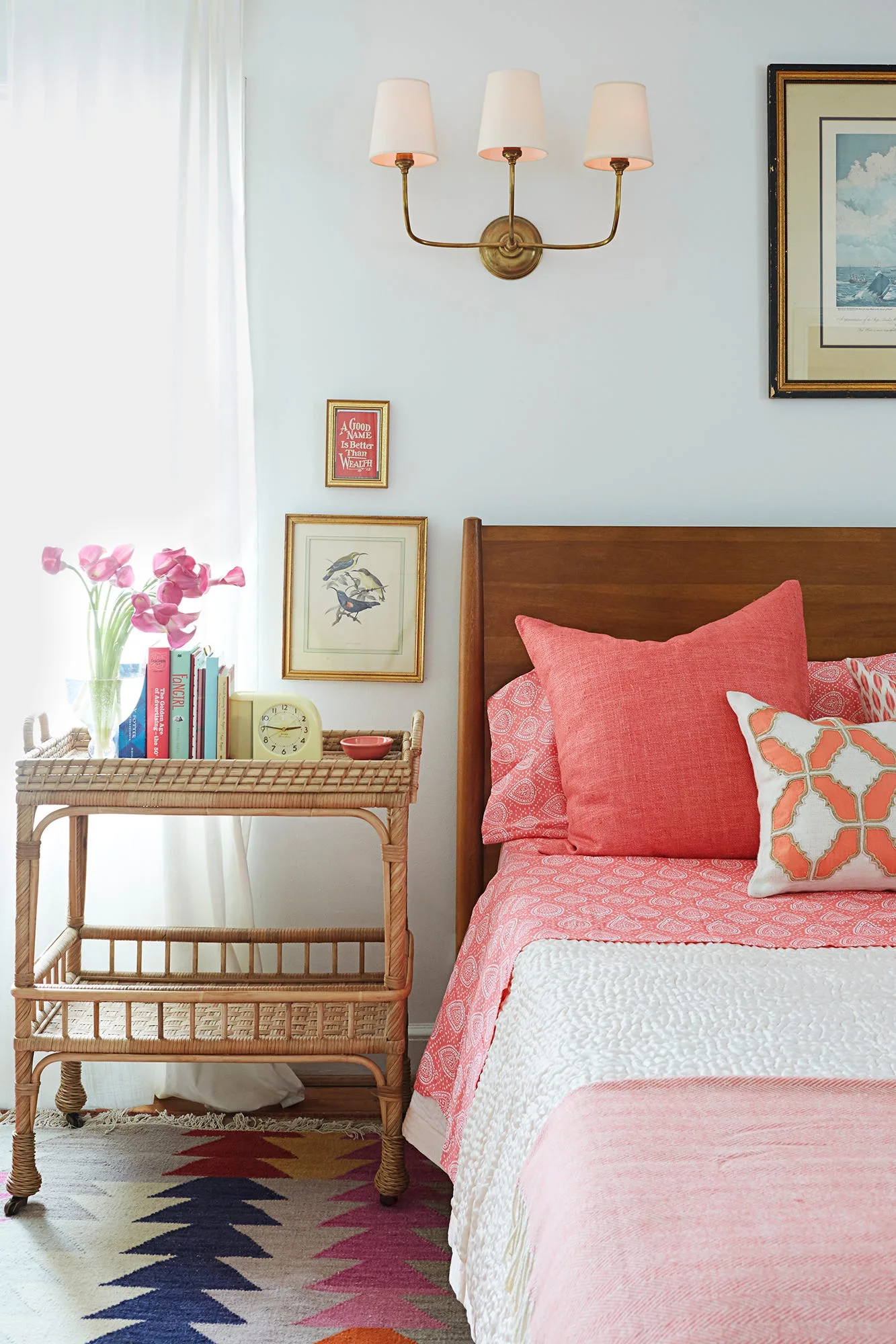 Bedroom with wooden headboard, pink bedding, woven two-tier trolley, framed bird pictures and multi-arm wall light beside pale curtains.