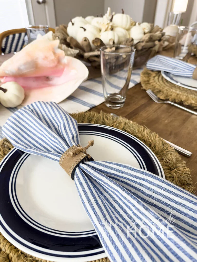 A blue-and-white striped napkin sits inside a wide brown ring above a navy-ringed white plate and fringed woven charger