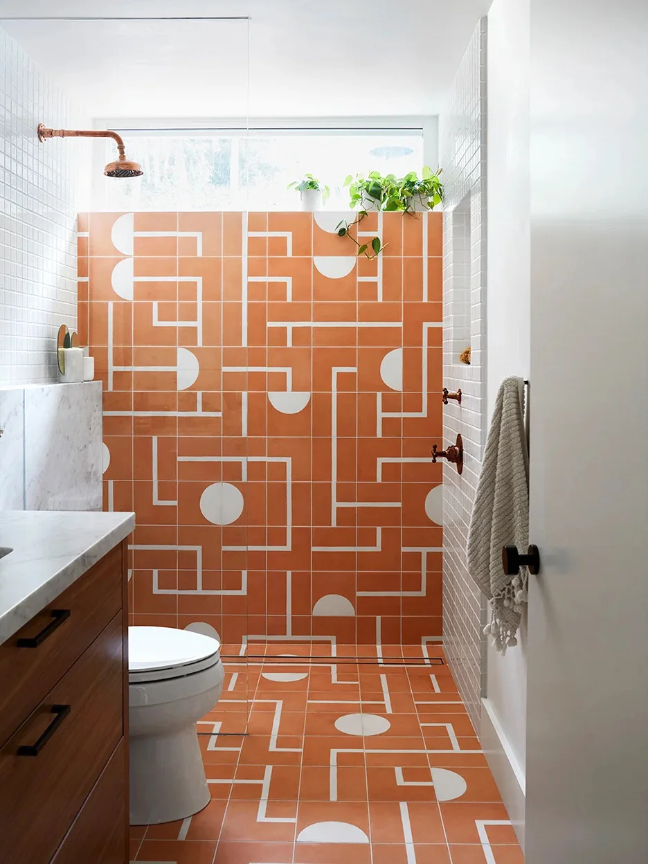 Orange geometric shower tiles repeat across a wall and floor beside white grid tiles, a wood vanity, a high window and a plant