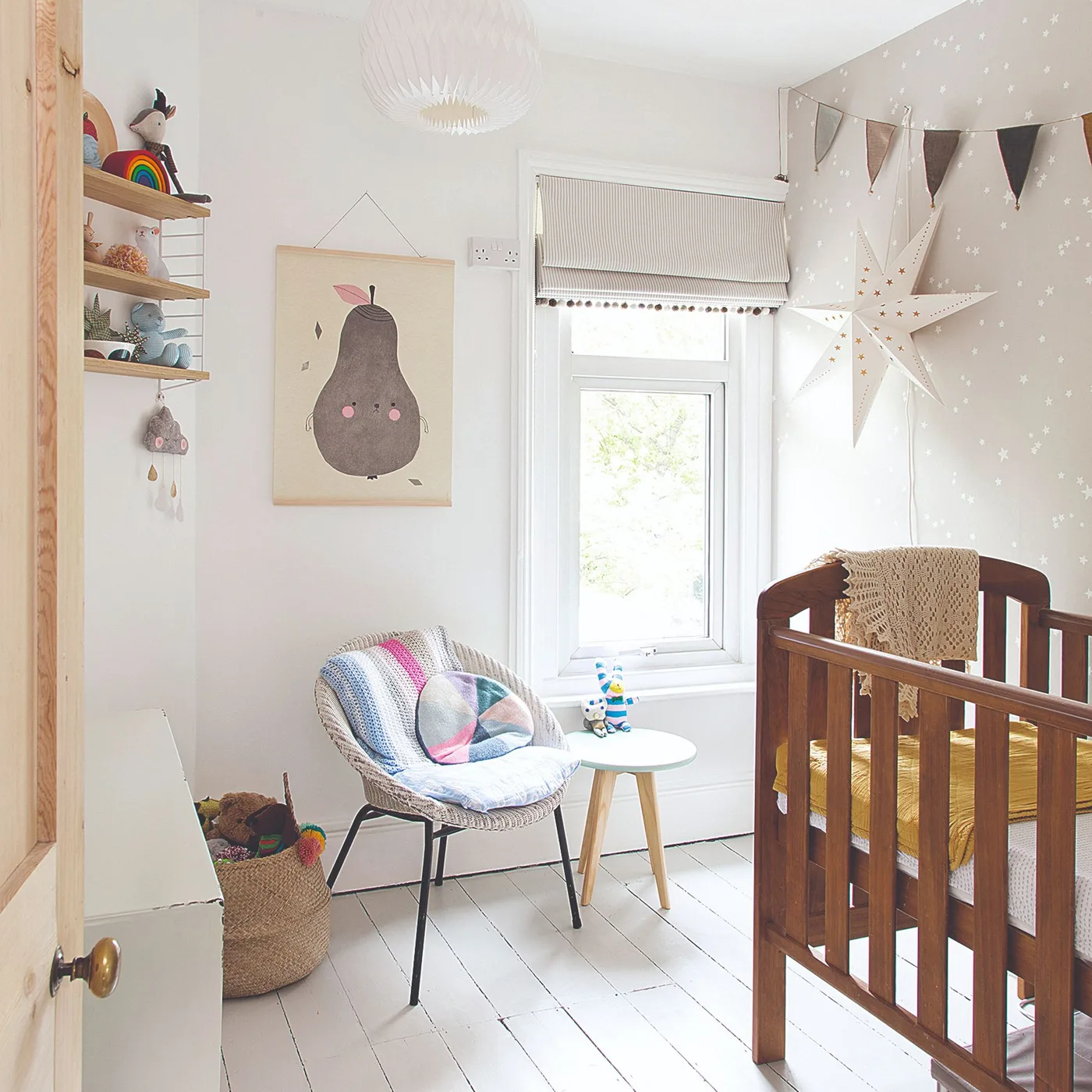Wooden crib in pale room with woven chair, basket, pear illustration, star-shaped wall piece and small side table.